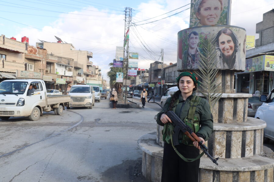 Das Bild zeigt eine junge Frau in der Uniform der kurdischen Frauenverteidigungseinheit YPJ. Sie trägt eine auf den Boden gerichtete Waffe in der Hand und lächelt in die Kamera