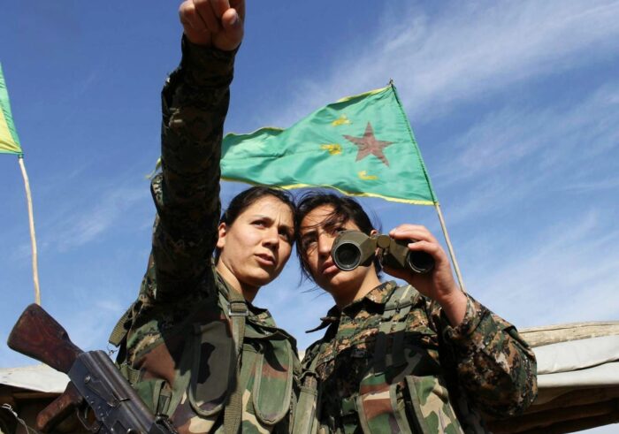 Zwei Frauen in den Uniformen der YPJ. Eine hält ein Fernglas in der Hand, die andere zeigt auf einen Punkt, weiter entfernt. Hinter ihnen weht eine Flagge der YPJ.