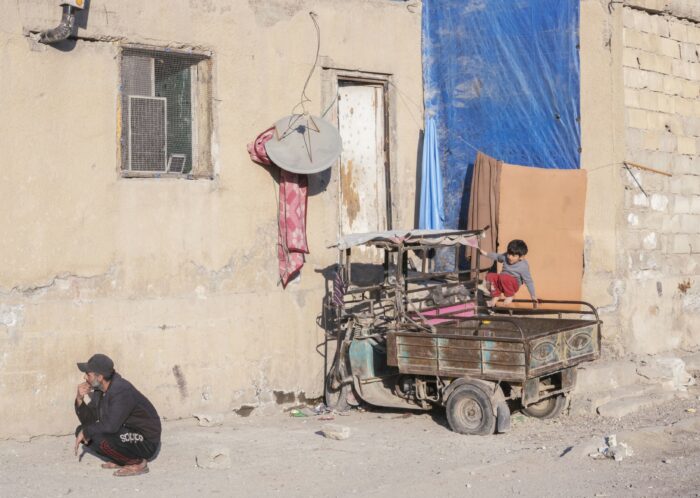 Eine Häuserwand, davor hockt ein Mann. Auf einem alten Anhänger, der neben dem Mann an der Wand steht, spielt ein kleines Kind