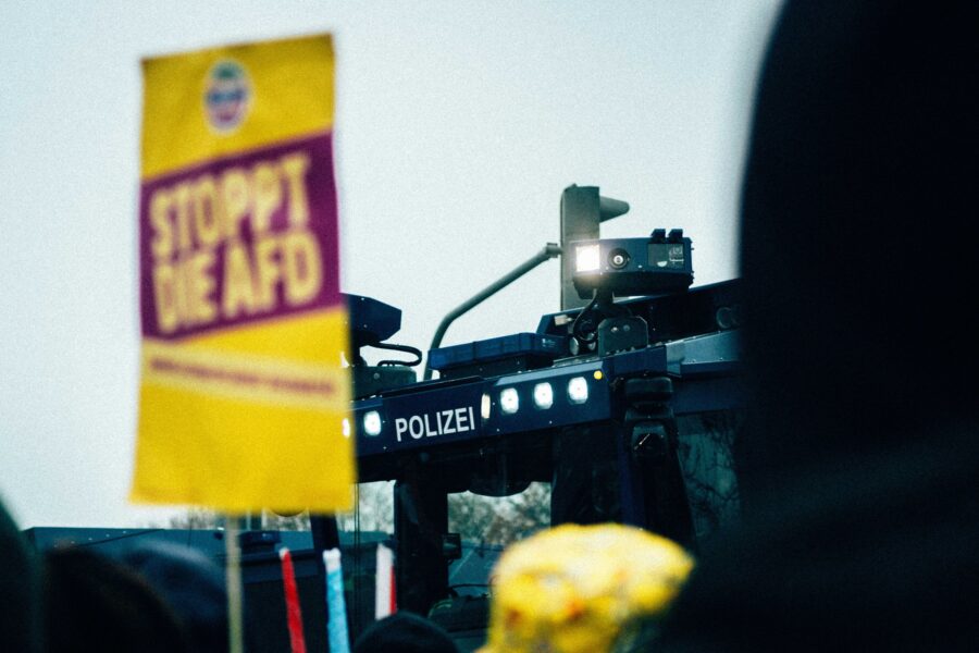 Demoschild mit der Aufschrift "Stoppt die AfD", dahinter ist ein Wasserwerfer der Polizei zu sehen