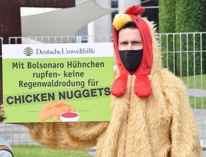 Ein als Huhn verkleideter Protestierender mit Schild in der Hand, auf dem steht: "Mit Bolsonaro Hühnchen rupfen. Keine Regenwaldrodung für Chicken Nuggets".