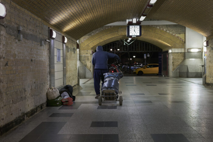 Man sieht eine U Bahn Unterführung und einen Menschen, der in der ecke liegt und einen, der daran vorbeigeht.