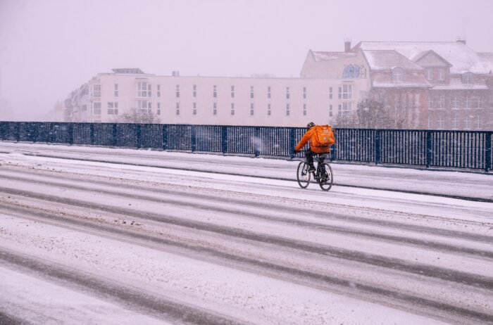 Eine verschneite Straße mit einem einsamen Fahrradkurier mit großer Tasche auf dem Rücken.