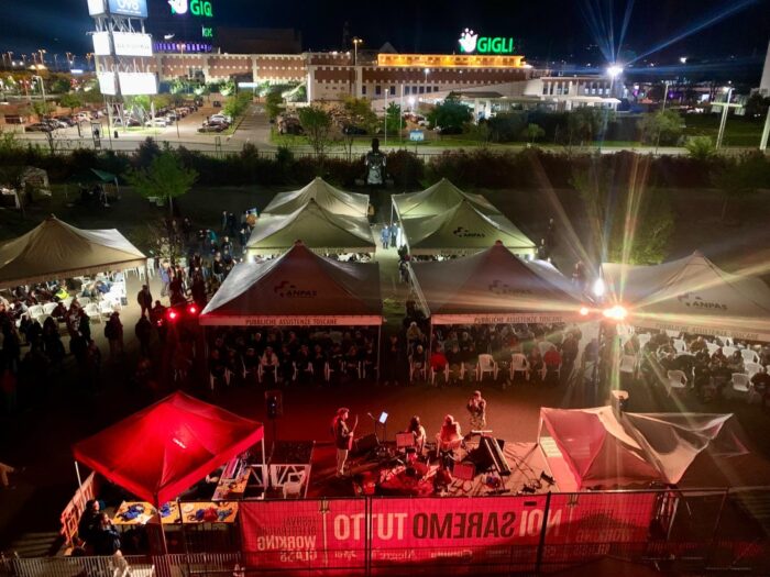 Ein Bühne in der Dämmerung mit Musiker*innen, davor Pavillion-Zelte mit Zuschauenden.