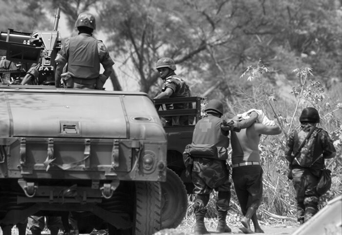 ein gepanzerter Fahrzeug, rechts davon mehrer Soldaten, die eine Person abführen