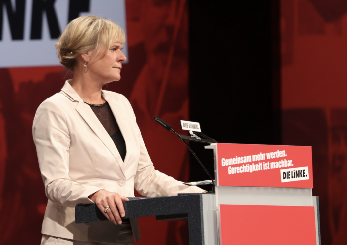 Man sieht eine Person in weißem Blazer an einem Redner*innenpult.