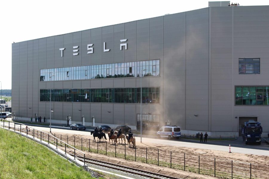 Große Halle mit dem Schriftzug Tesla, davor reiten Polizisten auf Pferden einen Sandweg entlang