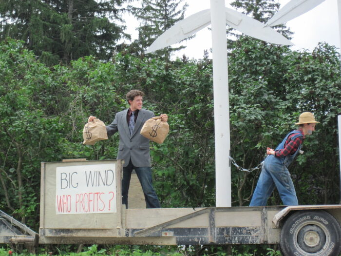Auf einer Bühne, ein Mann mit Geldsäcken in der Hand, in der Mitte eine Windkraftanlage, an der ein weitere Person gekettet ist.