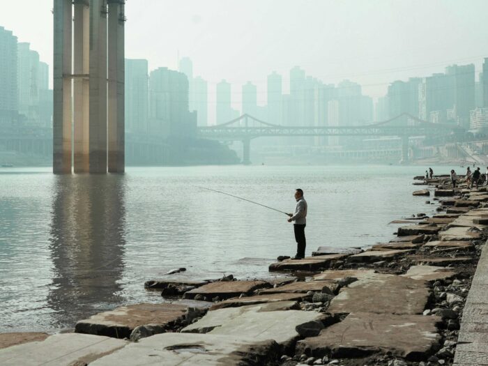 Ein Mann mit einer Angel steht an einem Fluss in einer Stadt, im HIntergrund ist eine Skyline aus Hochhäusern zu sehen, aus dem Fluss ragt ein riesiger Brückepfeiler auf.