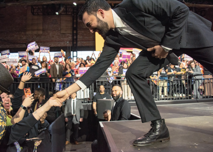 Man sieht Mamdani auf einer Bühne umringt von Fans, er reicht eine Hand nach unten in die Menge.