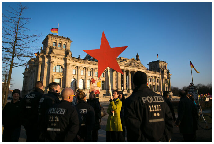 Demo gegen das PKK-Verbot vor dem Reichstag. Polizeibeamte stehen mit dem Rücken zur Kamera, in der Mitte hält eine Demonstrierende einen roten Stern hoch.
