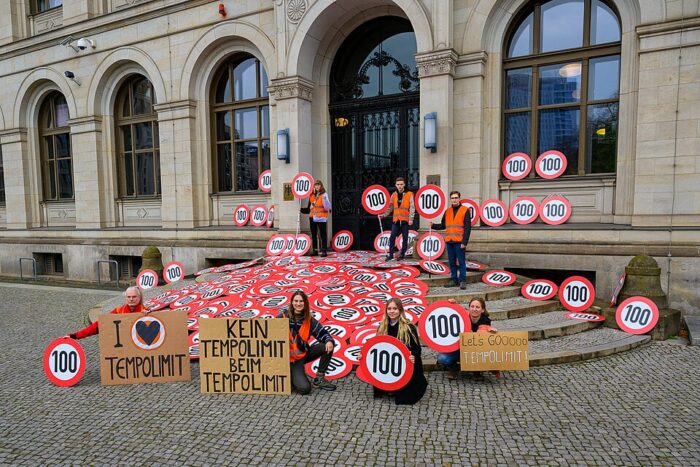 Eine Gruppe von Aktivist*innen vor und auf der Treppe eines Gebäudes. Auf den Treppen liegen ganz viele Verkehrsschilder mit der Aufschrift "100".