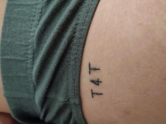 Ein "T4T"-Tattoo auf einem Bein unter dunkelgrünen Shorts