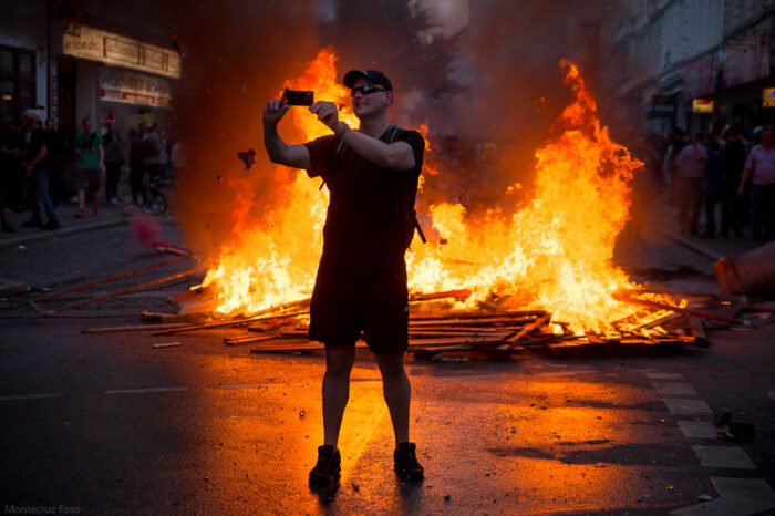 Ein Man mit kurzer Hose, Cap und Sonnenbrille macht ein Selfie vor einer brennenden Barrikade