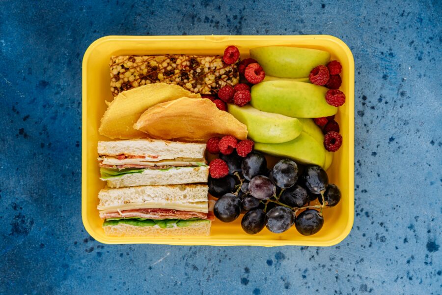 Man blickt von oben auf eine geöffnete Brotdose, die mit Obst und Broten gefüllt ist.