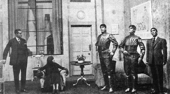Ausschnitt einer Szene aus einem Theaterstück. Die Bühne zeigt ein Wohnzimmer. Eine ältere Frau und ein Mann im Anzug begutachten Menschenähnliche Roboter.