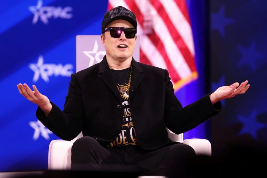 Elon Musk mit schwarzem Sakko, Sonnenbrille und Make-America-Great-Again-Cap sitzt mit ausgebreiteten Armen vor einer USA-Flagge