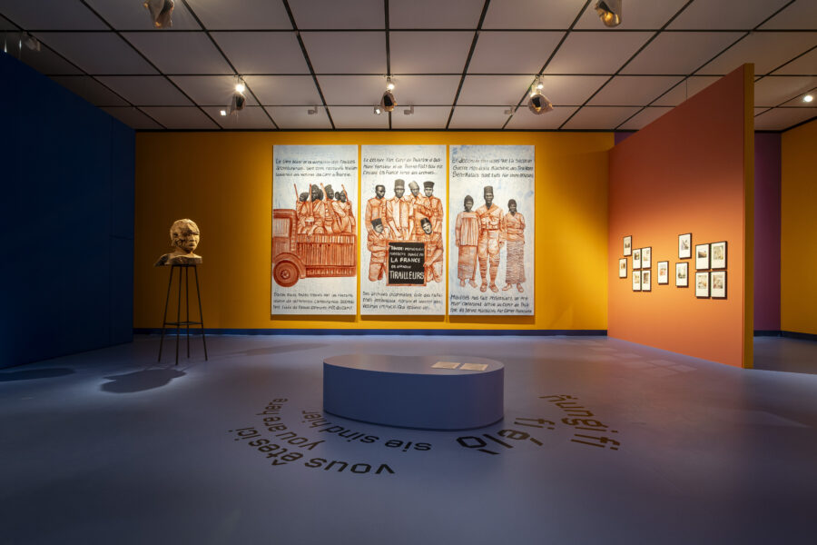 Foto einer Austellung mit Wänden in unterschiedlichen Farben. Zu sehen ist eine Skulptur und eine dreiteillige Illustration.