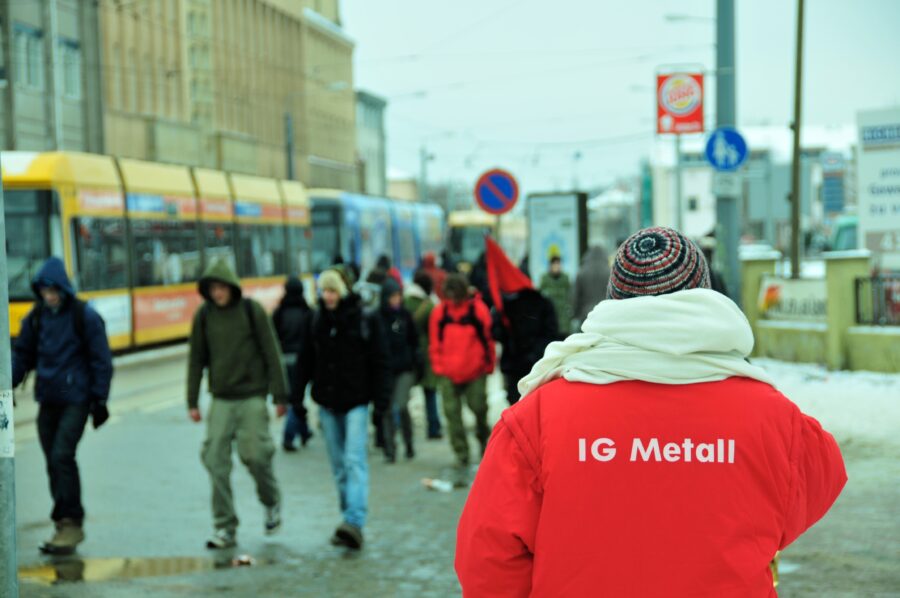 Man sieht an einer verregneten Straßenecke von hinten eine Person mit einer IG Metall Jacke.