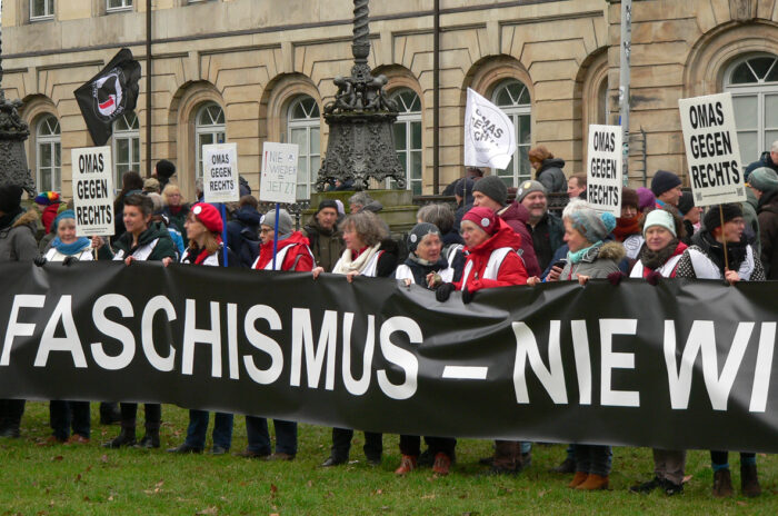 Eine Gruppe hinter einem langen Trasparent, dahinter Menschen mit Pappschildern