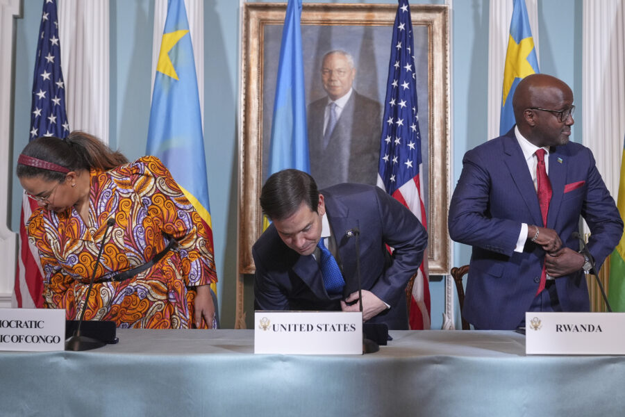 Drei offiziell gekleidete, zwei Männer und eine Frau, setzen sich an einem Tisch, hinter ihnen Flaggen von Kongo, Ruanda und den USA
