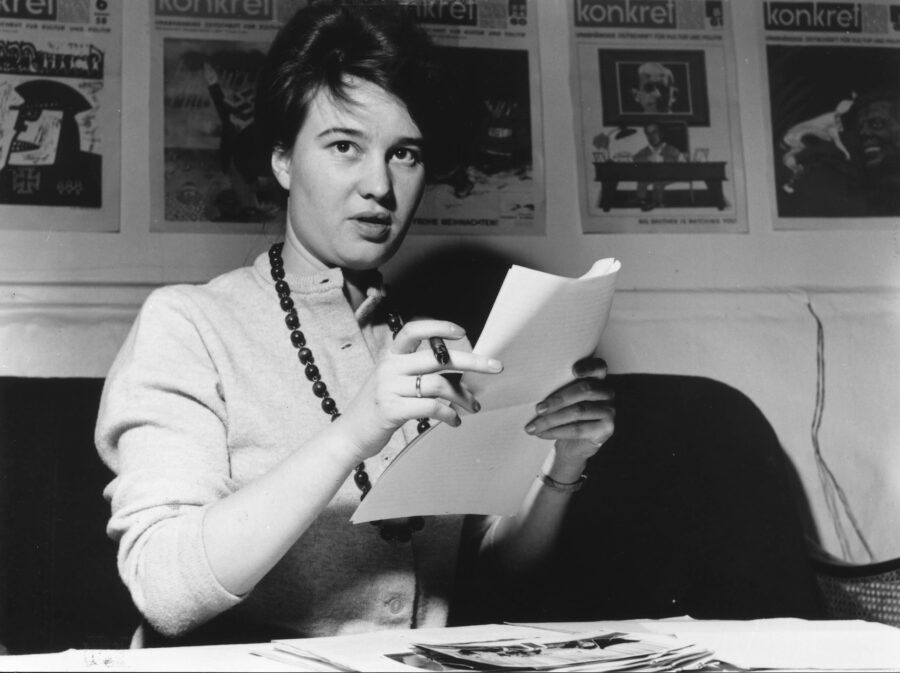 Ulrike Meinhof sitzt an einem Tisch in der konkret-Redaktion und hält ein Blatt Papier und einen Stift in der Hand