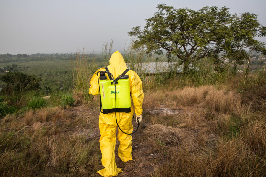 Eine Person in einem grellgelben Schutzanzug läuft alleine durch eine erhöhte Landschaft und sprüht eine Flüssigkeit auf den Boden