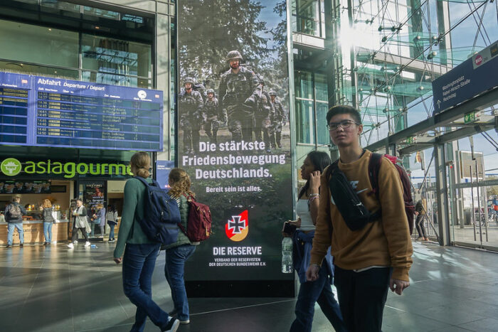 Eine Plakatwand in der Eingangshalle eines Bahnhofs, dahinter eine Fahrplananzeige, davor Menschen. Auf dem Plakat der Satz: "Die stärkste Friedensbewegung Deutschlands" auf dem Plakat Soldaten mit Waffe
