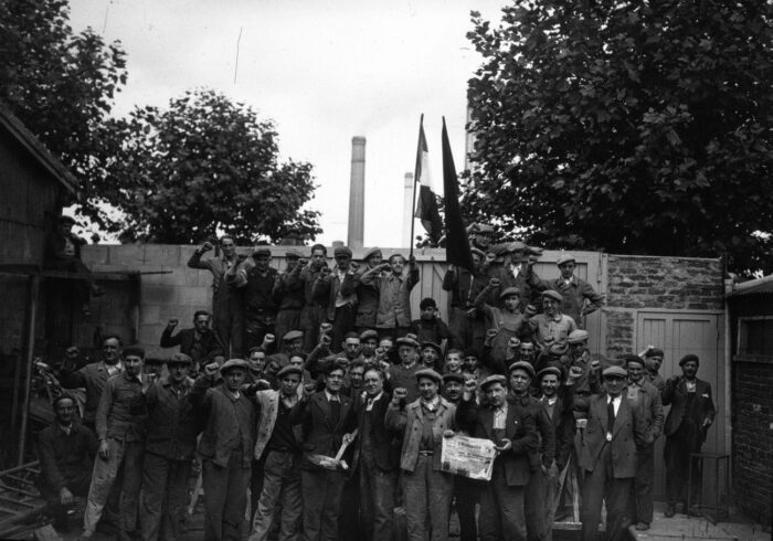 Geschätzt 40 bis 50 Arbeiter mit erhobenen Fäusten und kämpferischer Post vor einer Fabrik.