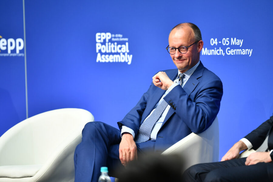 Das bild zeigt Bundeskanzler Friedrich Merz bei einer Veranstaltung der Europäischen Volkspartei. Merz sitzt auf einem Podium in einem weißen Ledersessel und grinst.