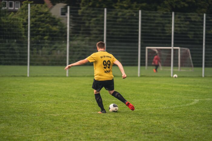 Das Bild zeigt einen Fußballer auf einem einfachen Rasenplatz, der auf ein Tor zielt. Er trägt ein gelbes Trikot mit der Aufschrift BV 09 Hamm