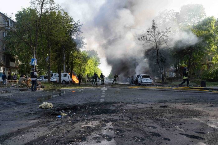 Straße mit einem Bombenkrater, im Hintergrund brennende Autos