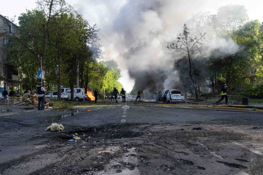 Straße mit einem Bombenkrater, im Hintergrund brennende Autos