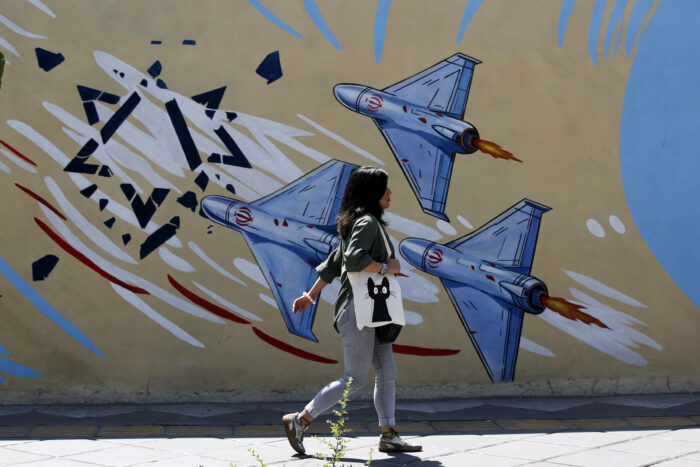 Eine Frau mit dunklen Haaren, heller Jeans und dunkler Bluse geht an einem Haus vorbei. Auf der Hauswand sind drei iranische Bomber zu sehen, die einen stilisierten Davidstern angreifen. Die Frau trägt auf ihrer rechten Schulter einen weißen Jutebeutel, auf dem eine schwarze Katze abgedruckt ist.