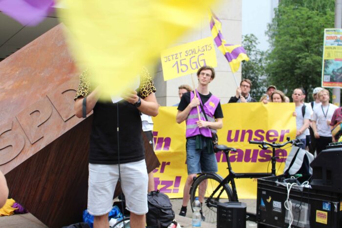 Demonstrant*innen in lila-gelber Ausstattung vor der SPD-Zentrale in Berlin. dem Redner hängt eine gelbe Fahne ins Gesicht, links neben ihm ein ist ein Metallblock mit SPD-Aufschrift, dahinter steht ein Demonstrant mit einem Schild mit der Aufschrift: Jetzt schlägt's 15 GG