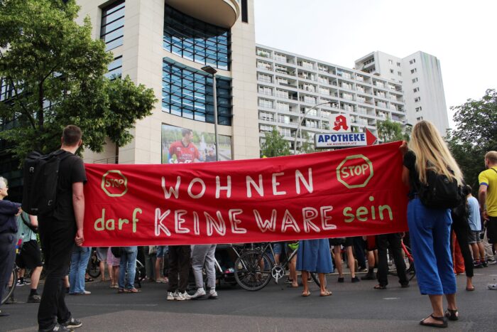 Man sieht Menschen, die ein rotes großes Transparent hochhalten, auf dem steht: Wohnen darf keine Ware sein.