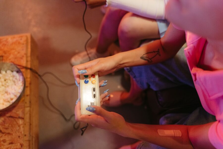 Eine Person in einem pinken Shirt hält einen Controller und sitzt an einem Tisch mit Popcorn. Daneben sitzt eine zweite Person.
