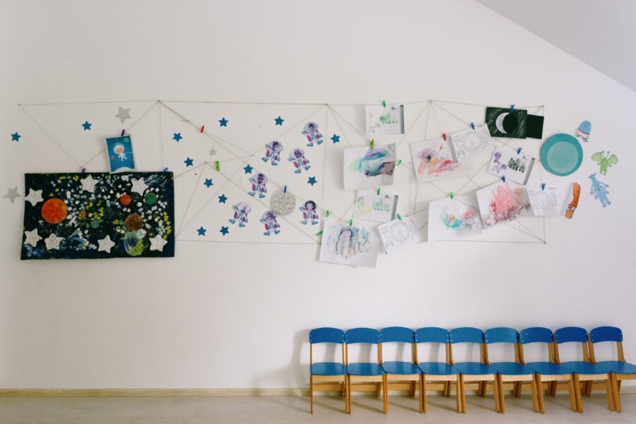 Das Bild zeigt eine Reihe Kinderstühle vor einer Wand mit vielen bunten, selbstgemalten Bildern