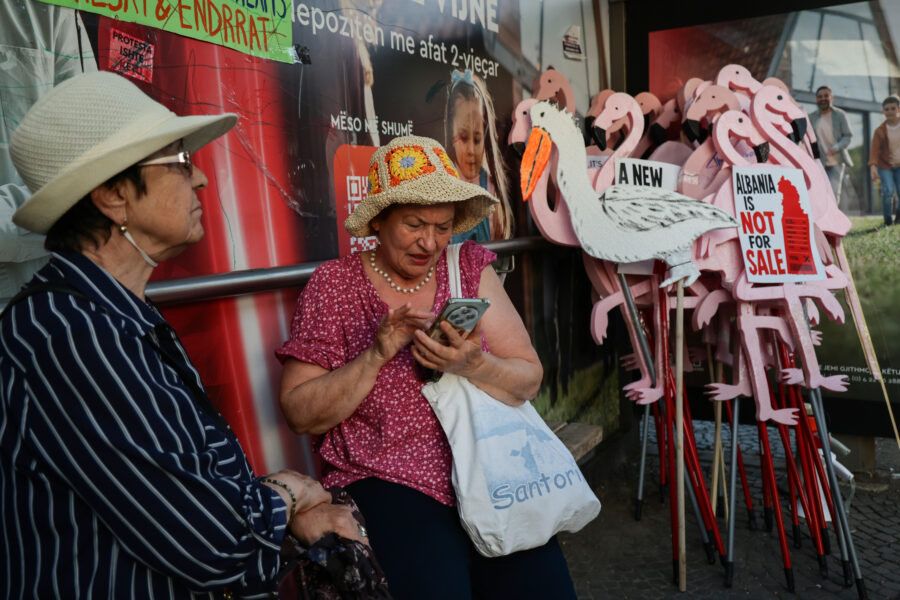 Zwei ältere Frauen sitzen an einer Bushaltestelle. Hinter ihnen in der Ecke sind Protestschilder in Form von Flamingos