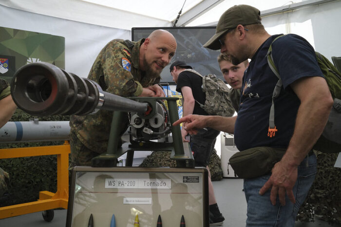 Ein Soldat beugt über ein schweres Geschütz und schaut dabei einen Mann an, der sich interessiert die Waffe anschaut. Im Hintergrund weitere Menschen und die Innenwand eines Zeltes