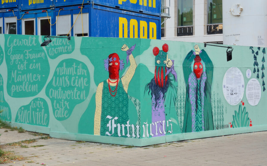 Man sieht eine Mauer mit einem Graffiti mit drei vermummten Personen, die kämpferische Gesten machen.