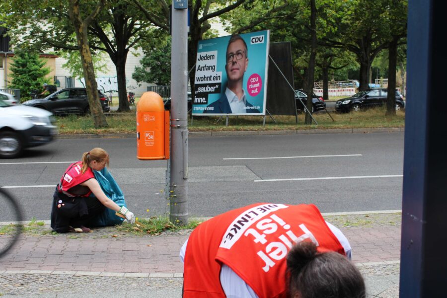 Linke-Mitglieder in roten Westen sammeln Müll, im Hintergrund ist ein Großplakat von CDU-Kandidat Stefan Evers zu sehen, auf dem steht: Jedem Anfang wohnt ein "sauber" inne.