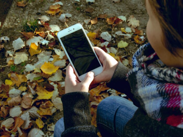 Man sieht Herbstlaub und eine sitzende Person, die auf ein Handy schaut.