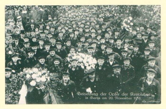 Man sieht eine alte schwarzweiß Postkarte, die wiederum eine Beerdigung von Revolutionär*innen im Jahr 1918 zeigt.