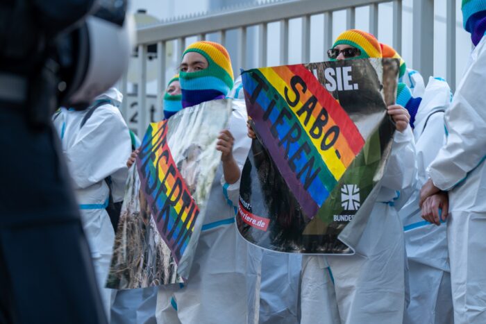 Vor einem Zaun sind Demonstrierende in weißen Overalls mit Protestplakaten zu sehen.