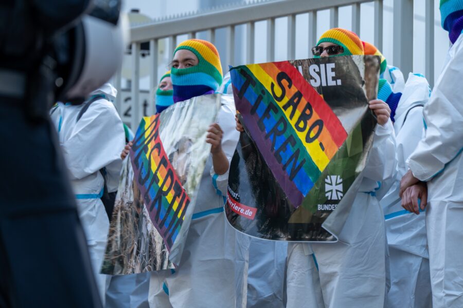 Vor einem Zaun sind Demonstrierende in weißen Overalls mit Protestplakaten zu sehen.