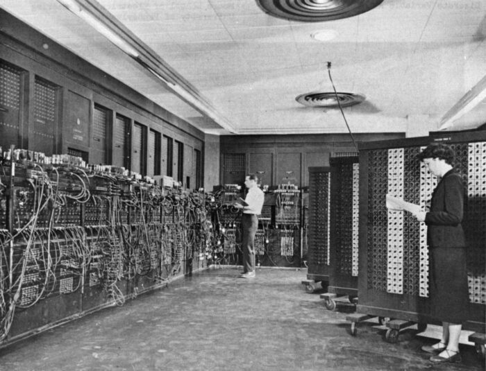 Die Programmiererin Betty Holberton und der Programmierer Glen Beck stehen mit Klemmbrettern vor vor wandgroßen Computer.