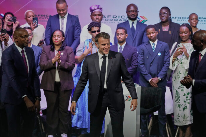 Mehrere Menschen stehen in zwei Reihen für ein Foto, vor ihnen steht Macron in einer komischen Pose.