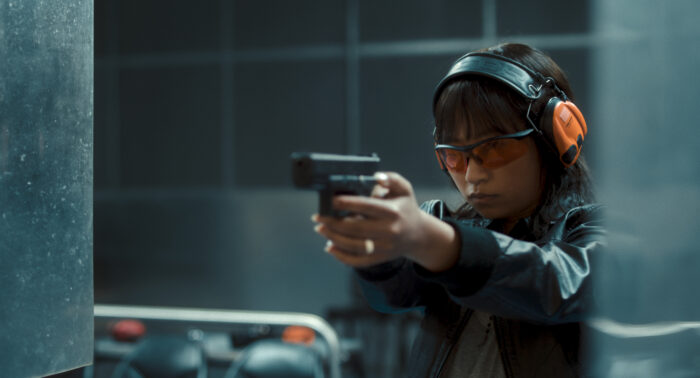 Filmstill aus dem Film "Staatsschutz", das die Protagonistin Seyo Kim zeigt, wie sie das Schießen übt. Sie trägt eine Schutzbrille, eine Lederjacke und Ohrenschutz.