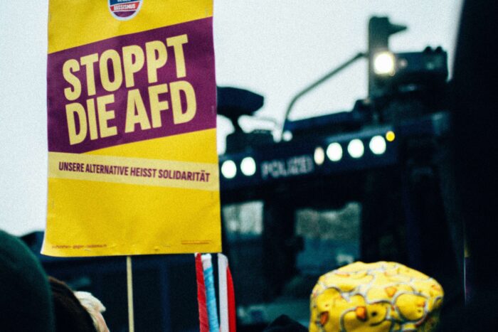 Gelbes Schild mit der Aufschrift "Stoppt die AfD", dahinter ein Polizeiwasserwerfer